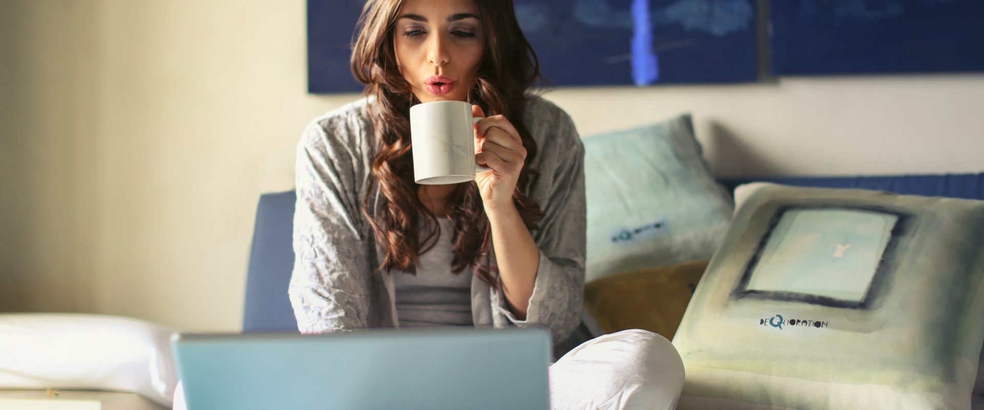 a woma sitting on her bed sipping on tea and scrolling on her laptop